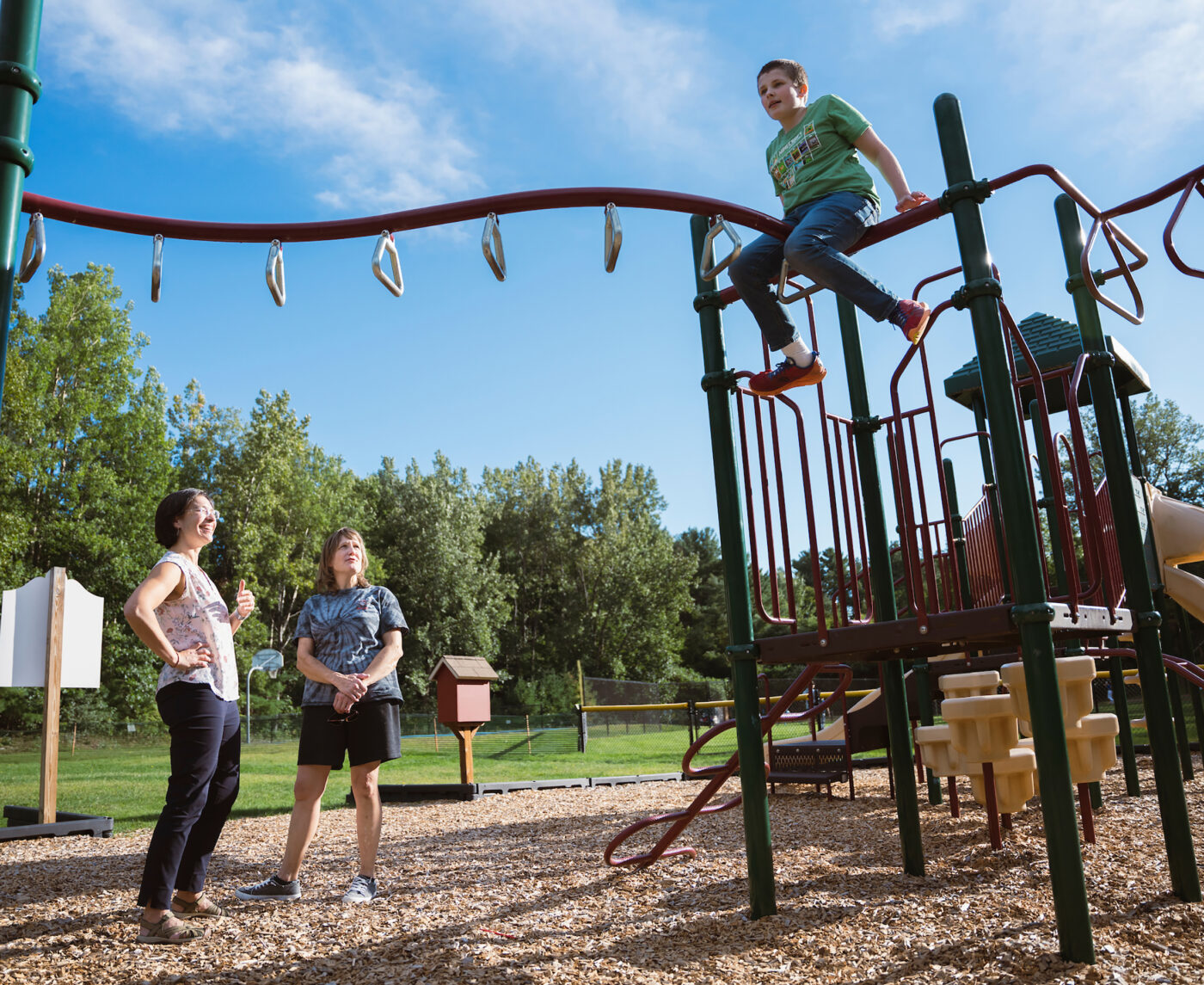 A pale-skinned boy sits atop a jungle gym as two middle-aged white women look on