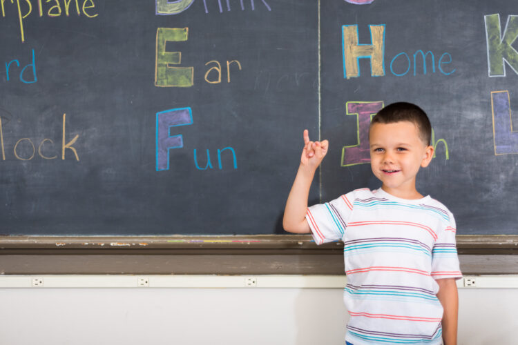 A young pale-skinned boy holds his hand up to sign the letter "i" in ASL, smiling shyly