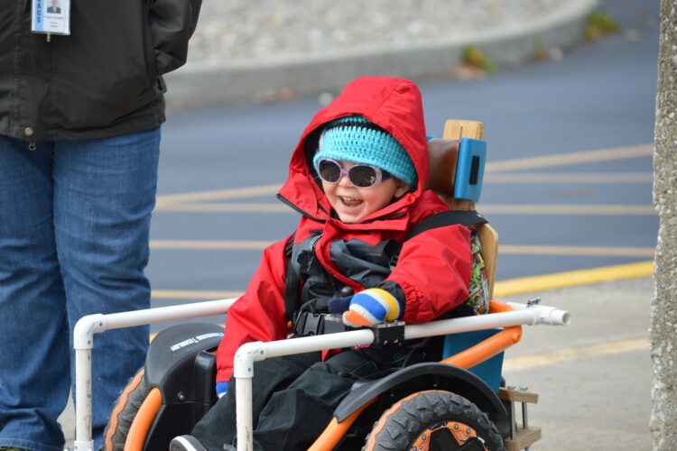 Young child wearing red winter coat and sunglasses controls a powered wheel chair with adapted PVC pipe frame around it.