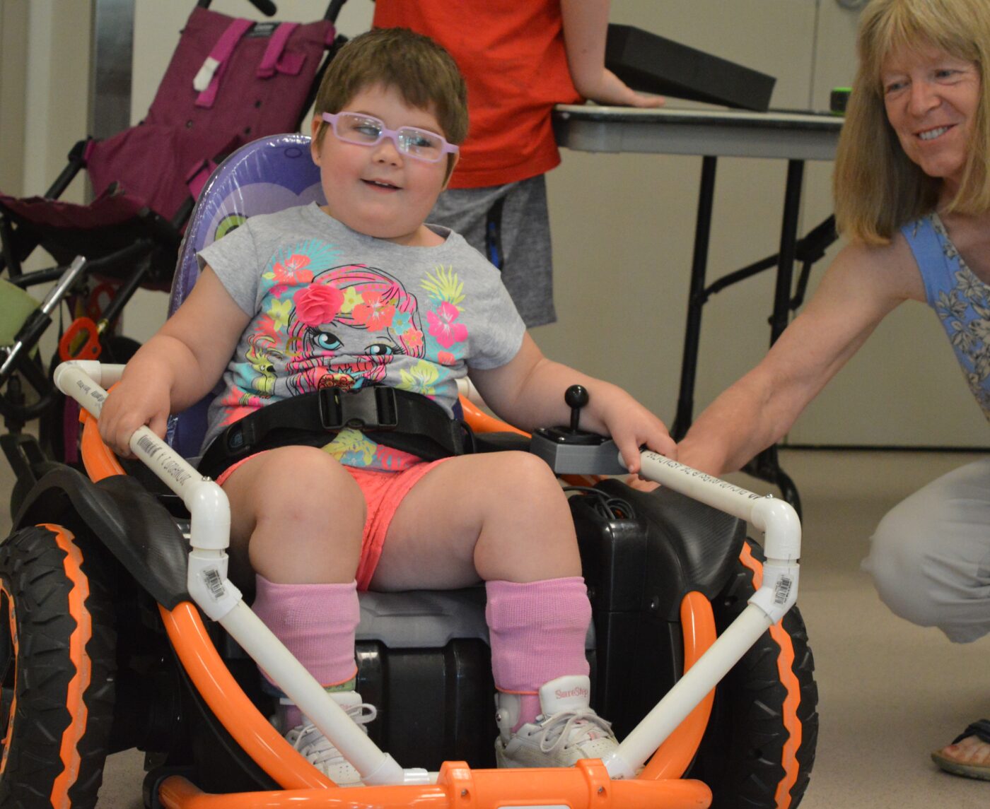 A small child with glasses is strapped into a motorized chair as a woman kneels nearby