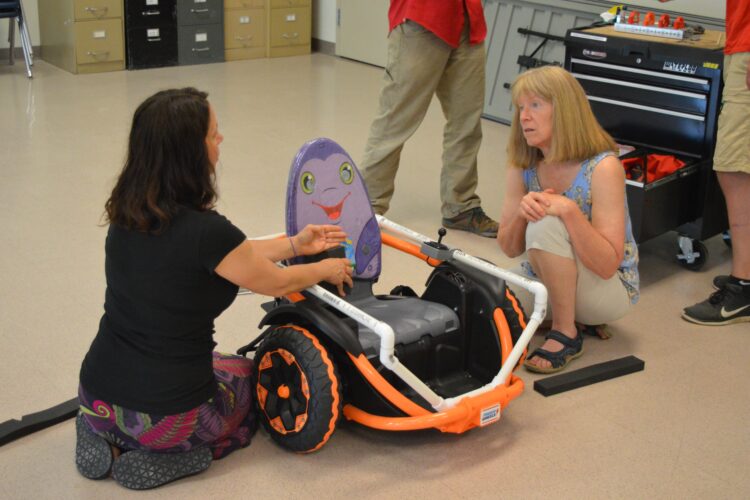 A middle-aged woman kneels beside a very small motorized wheelchair, showing it to another woman crouched nearby