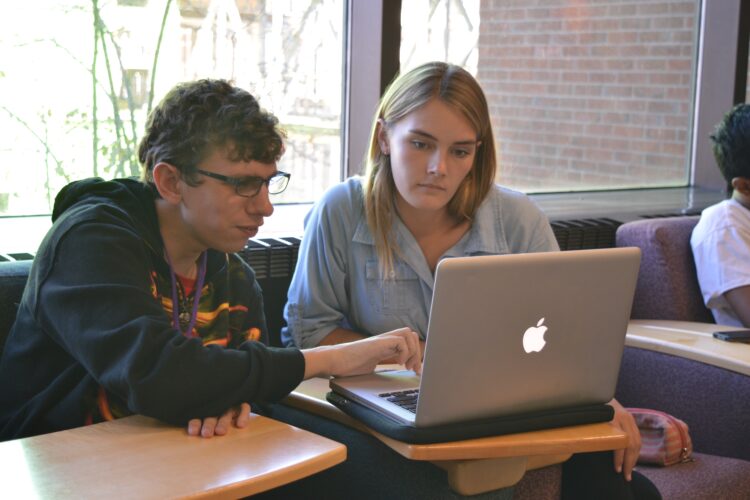 A college-aged man with short curly hair and glasses points to something on a laptop, while seated next to a college-aged woman with blonde hair, who is looking at the screen