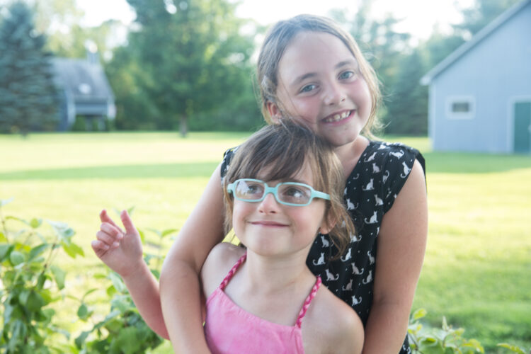 A small child with blue glasses is hugged by a larger girl smiling shyly, in front of a vast green lawn