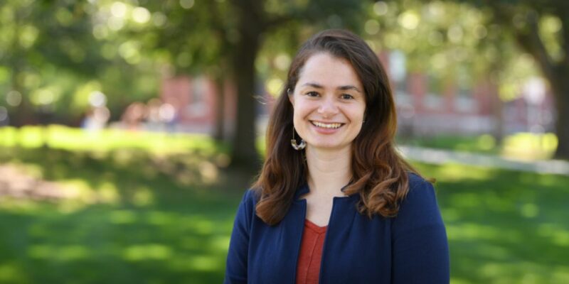 A pale-skinned woman in her mid-thirties, with thick walnut-brown hair worn loose to her shoulders smiles happily at the camera