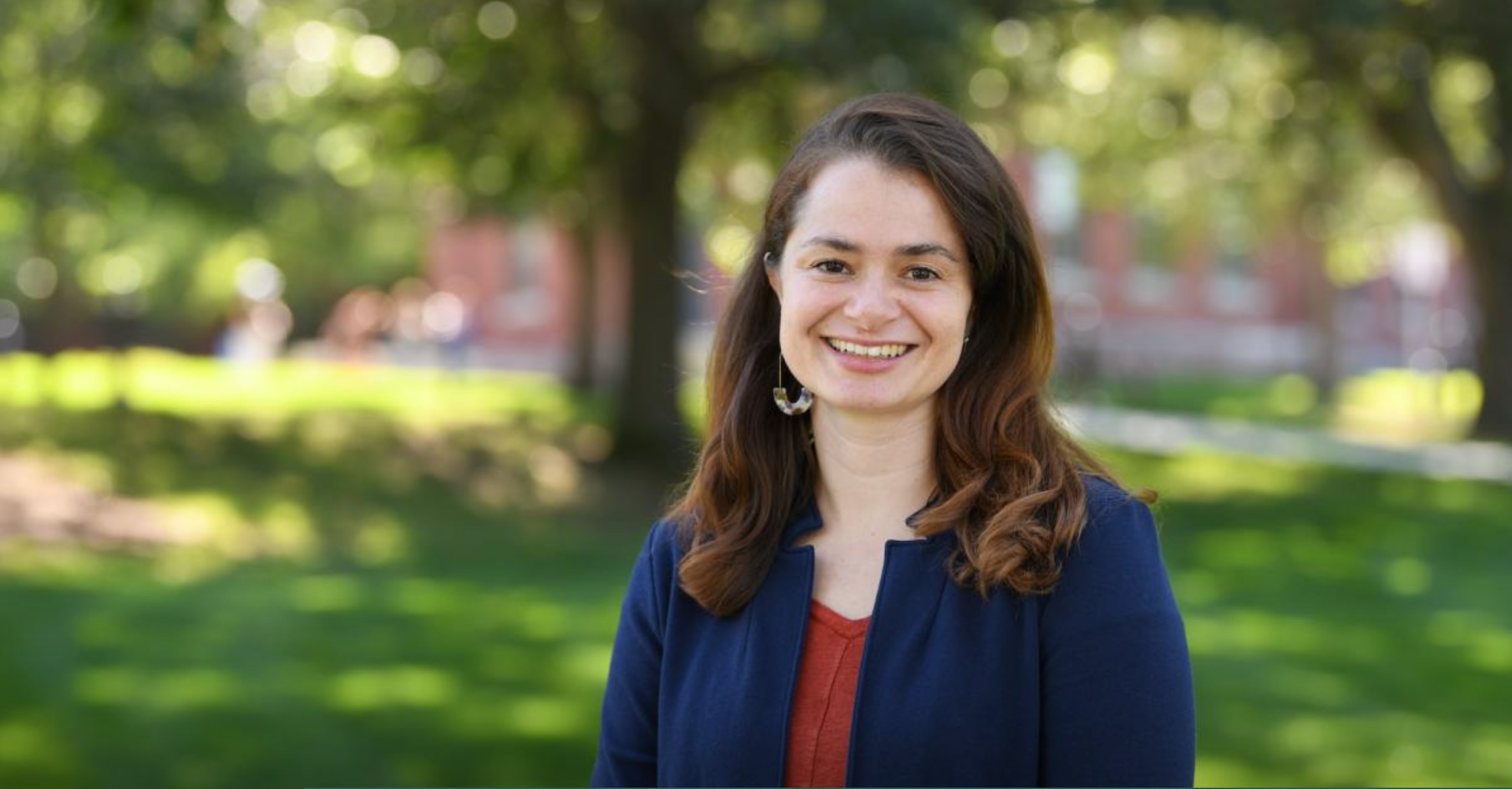 A pale-skinned woman in her mid-thirties, with thick walnut-brown hair worn loose to her shoulders smiles happily at the camera