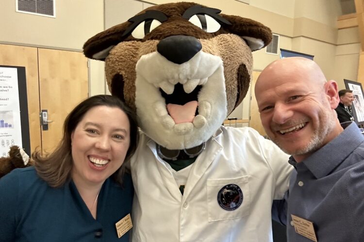 A pair of middle-aged pale-skinned people stand either side of a UVM catamounts mascot in a lab coat