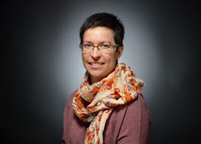A pale-skinned woman in her mid-forties with short dark hair and thin-rimmed glasses, wearing a colorful floral scarf