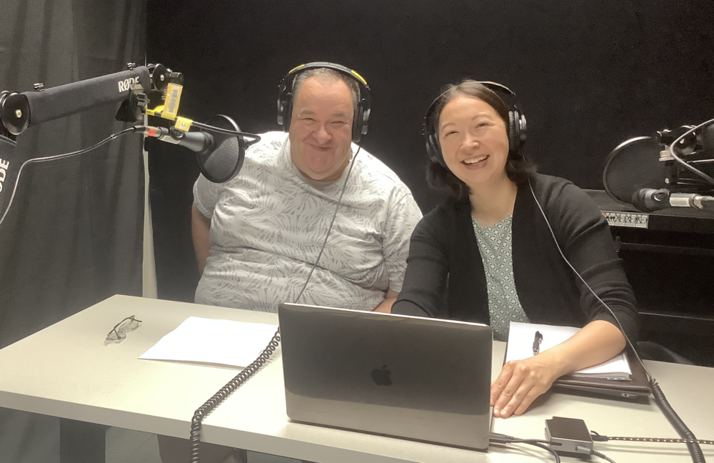 A full-figured white man in his sixties sits next to a mixed-race Asian woman in her forties at a table. Both are wearing headphones and a microphone is in front of the man