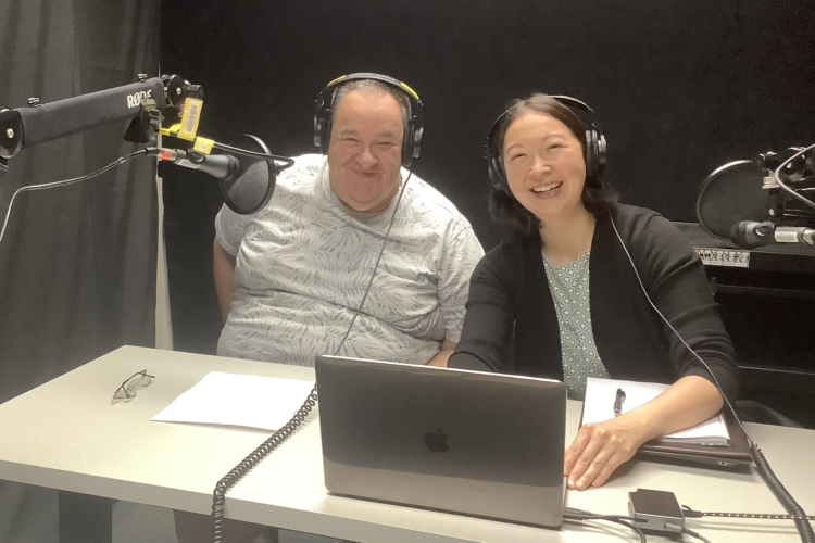 A full-figured white man in his sixties sits next to a mixed-race Asian woman in her forties at a table. Both are wearing headphones and a microphone is in front of the man