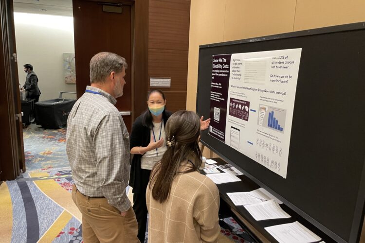 A mixed-race Asian woman in her forties explains a research poster to two people at a conference