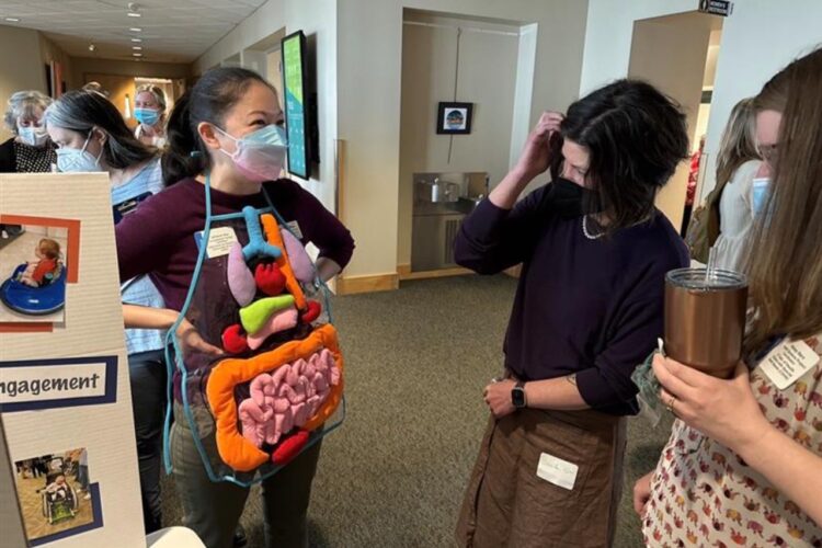 A masked, mixed-race Asian woman in her forties models an anatomical apron showing the digestive system for a curious onlooker at a conference.