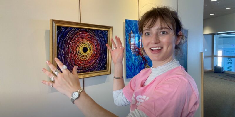 A pale-skinned college-aged woman in a bubblegum pink tee with a white turtleneck underneath expresses joy while hanging a painting of a black hole surrounded by colors