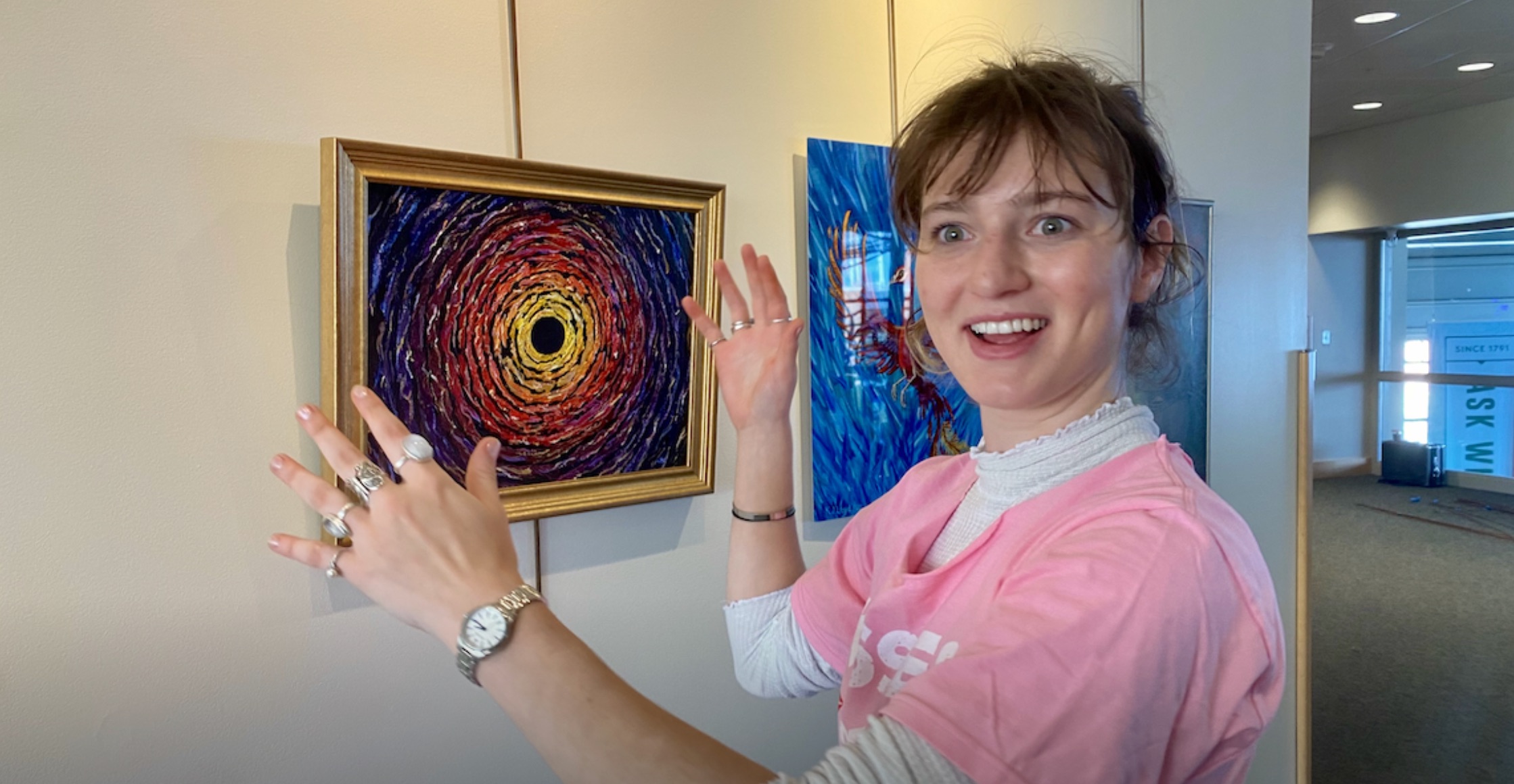 A pale-skinned college-aged woman in a bubblegum pink tee with a white turtleneck underneath expresses joy while hanging a painting of a black hole surrounded by colors