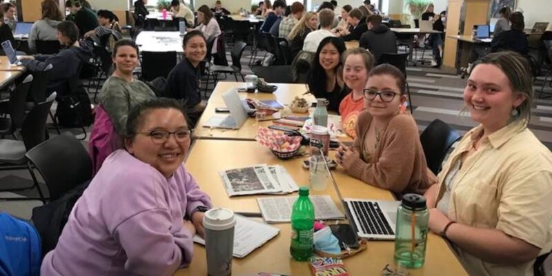 A group of college students, some with visible disabilities, some without, look up at the camera from where they're gathered around a cafeteria table