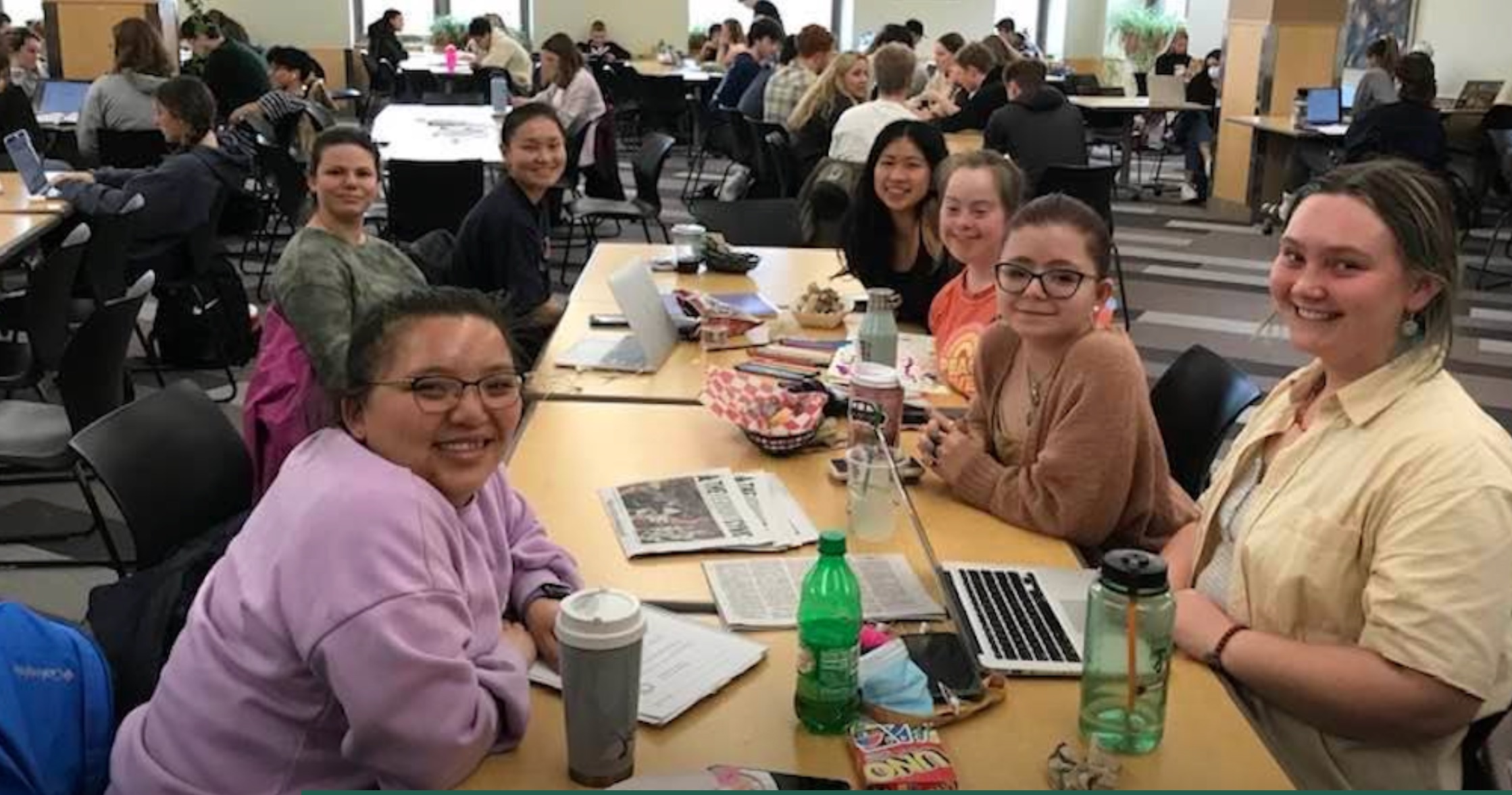 A group of college students, some with visible disabilities, some without, look up at the camera from where they're gathered around a cafeteria table