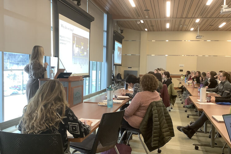 A blonde woman in her mid-fifties addresses a packed classroom