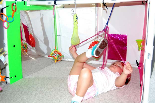 Figure 11a. A toddler is lying on a rug on her back in a “Little Room,” under a variety of
interesting objects that are hanging above her. She is holding a bright pink curly string of
material in each hand, and exploring a third string of material with her right foot. She is
also looking at the bright pink color. Possible concepts being developed are: “The world
around me is interesting and fun. I like things that are brightly colored. I like things that
make noise. I can explore by myself.”