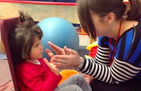 Figure 2a. A young girl sits on the floor in a supported seat, facing her intervener. She
and the intervener have their hands up in the air, ready to play a Pat-a-Cake game. She
is making eye contact with her intervener.