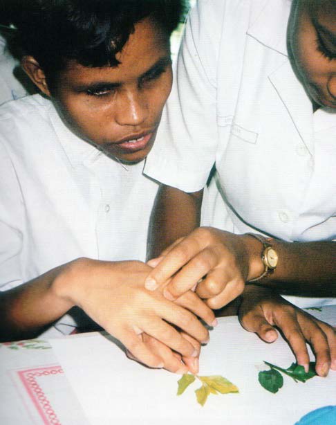 A dark-skinned DeafBlind young woman with short wavy black hair moves her hand over a book with leaves on the pages, as an older woman puts her hands over the top of the DeafBlind woman's.