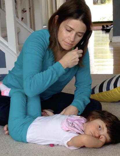 A pale-skinned DeafBlind toddler girl lies on a carpet as a woman brushes her own dark hair, while leaning over the girl and touching the girl's hands