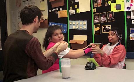 An almond-skinned DeafBlind adolescent girl sits with an adult and a Black female peer at a table. The peer holds the DeafBlind girl's hand as the adult signs against the DeafBlind girl's shoulder.