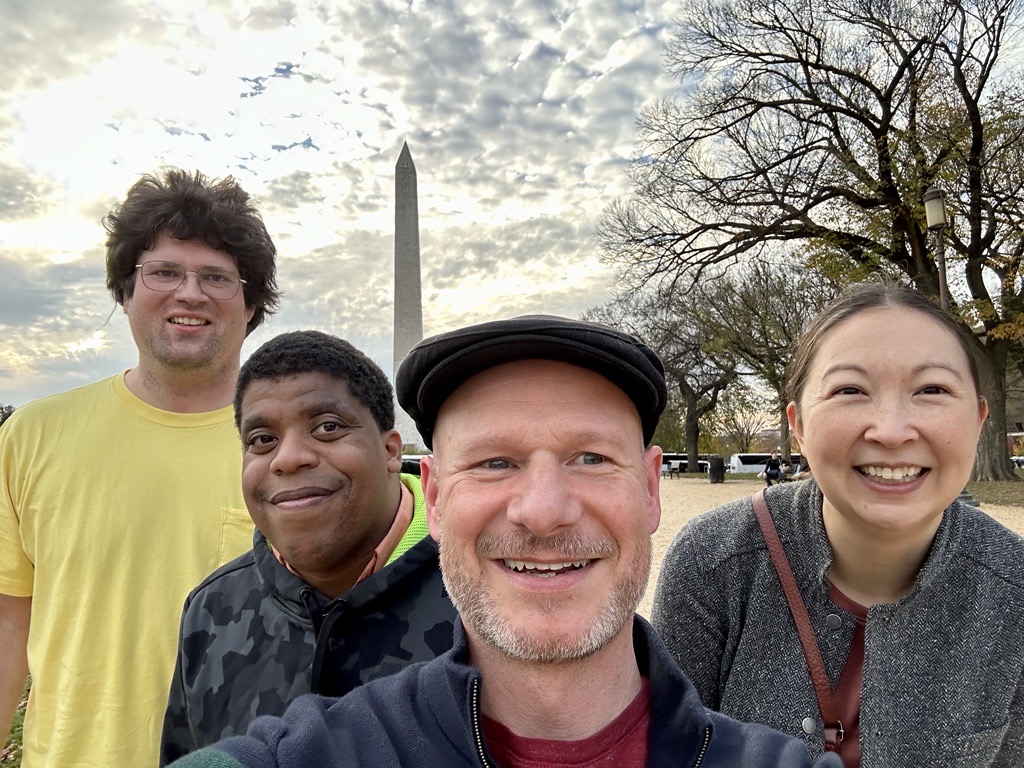 A bald white man in his fifties holds the camera for a selfie with a white man with brown curly hair, a Black man, and a mixed-race Asian woman in her forties, with the Washington monument in the background