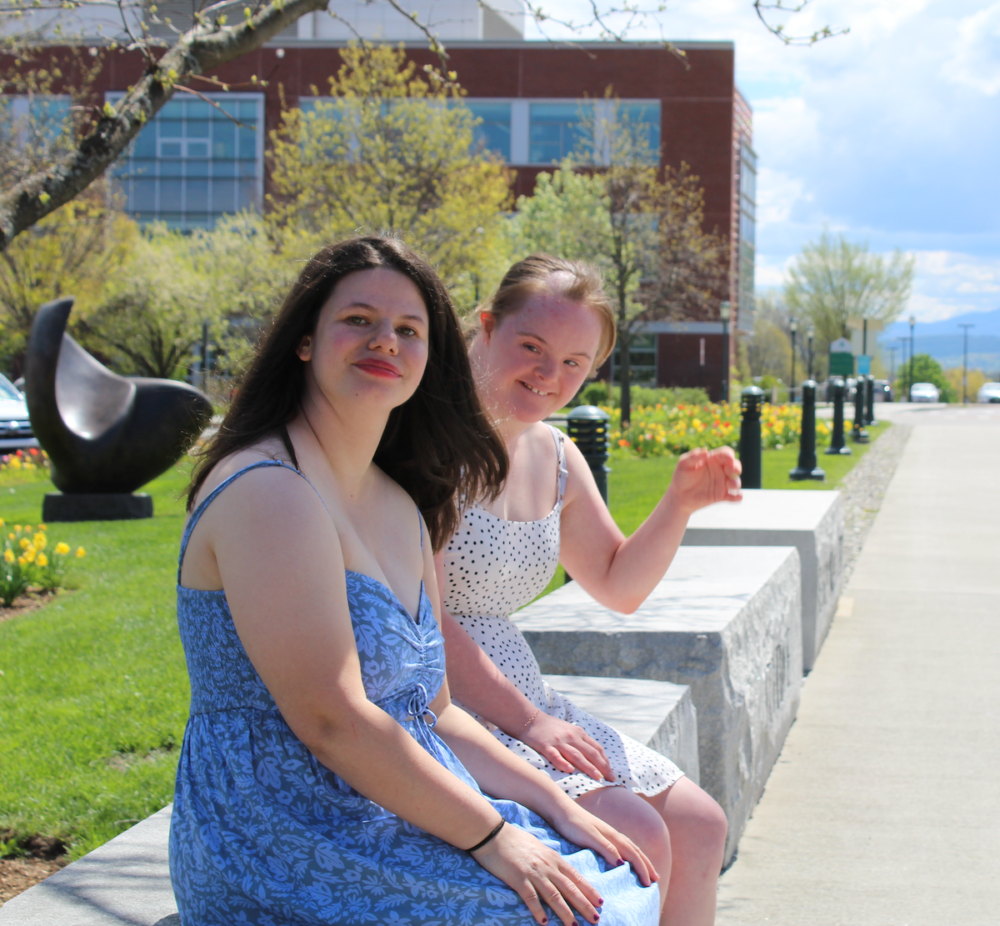 Two college-aged women in sundresses, one with dark, loose hair and almond skin, the other with lighter hair and Down Syndrome, sit in the sunshine on a green college campus. The woman with Down Syndrome waves at the camera