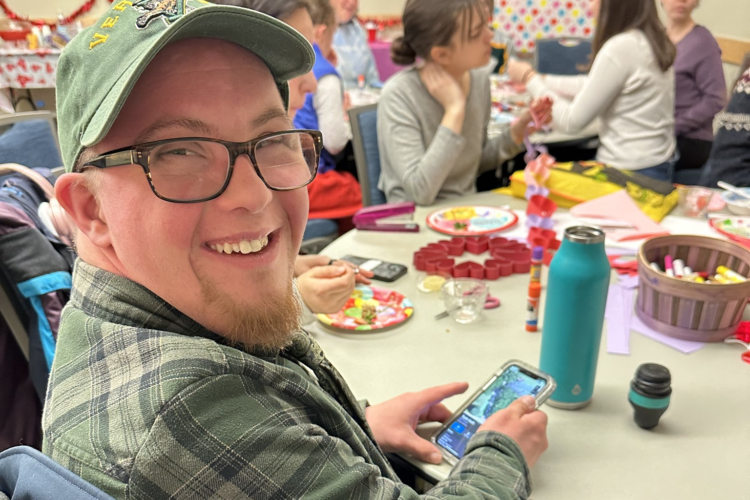 A college-aged man with Down syndrome, wearing glasses, a Vermont ball cap and a green flannel shirt, smiles over his shoulder at the camera. Behind him, a Valentines party is in full swing.