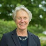 Katharine Shepherd: a pale-skinned woman in her mid-sixties, with short white hair in a pageboy, wearing a dark blue suit jacket and linked circle rings necklace, smiling broadly at the camera