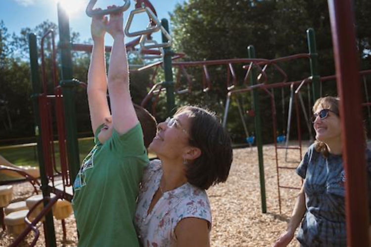 A middle-aged woman with short dark hair supports a pre-teen boy as he hangs from the monkey bars, while another woman looks on