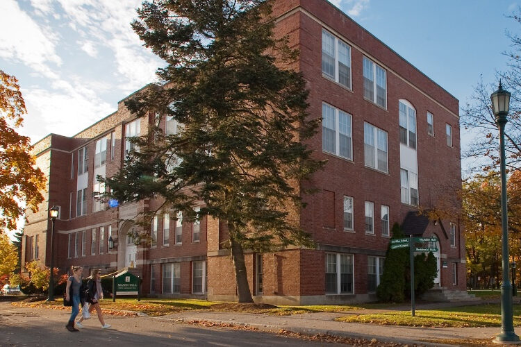 a three-story red brick building on a sunlit campus corner, near a crossroads sign. Two college undergrads walk past.