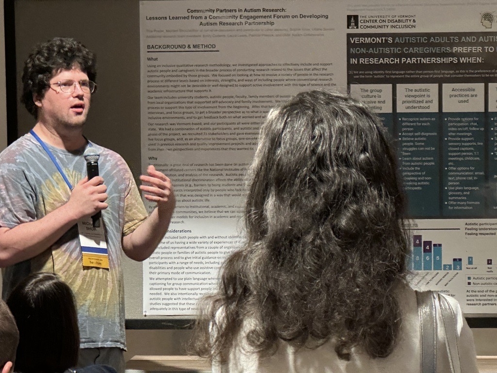 A white man in his thirties, with dark curly hair, speaks into a microphone next to a research poster mounted on a wall, as an audience listens