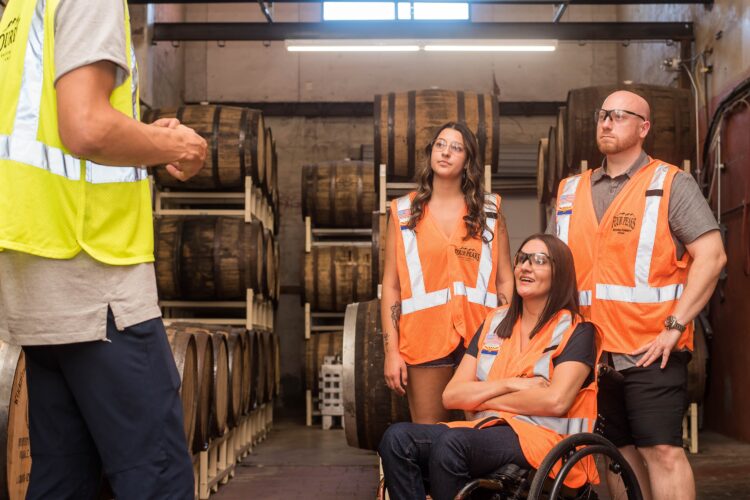 Three people in orange reflective safety vests, one in a wheelchair, listen to a faceless person in a yellow safety jacket in an industrial workplace