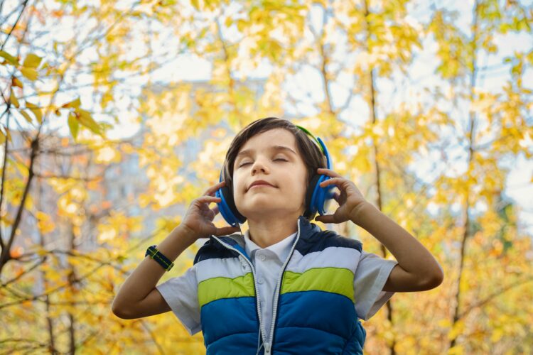 A small boy holds headphones over his ears and closes his eyes in the middle of a forest grove