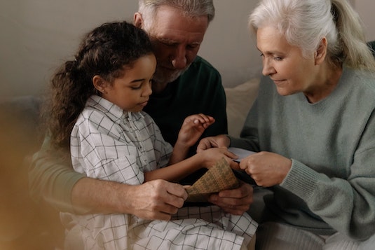 A dark-skinned girl sits on the lap of an older, white-haired man as a woman next to him tends to the girl's hands.