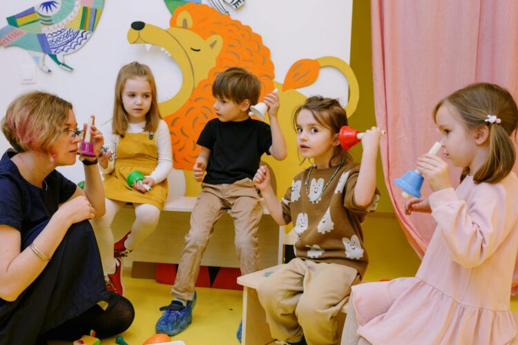 A group of children site in a circle playing with plastic colored bells as a teacher looks on. One little girl holds the bell to her ear while looking directly in the camera.