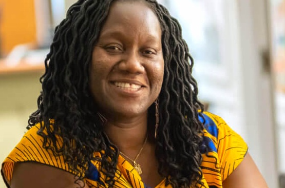 A Black woman in her forties with hair in loose, curled locs to her shoulders sits smiling, in a gold and cobalt short-sleeved top