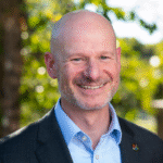 Jesse Suter, smiling white 50 year old bald man with short grey facial hair, wearing a grey jacket and blue button up shirt.
