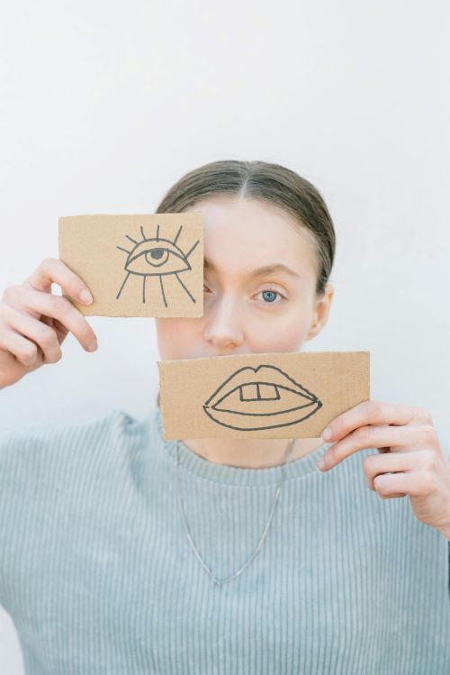 Young person holding in light blue sweater sitting at table holding cardboard pieces with face drawings over their own face